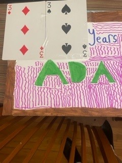 Green ADA lettering with pink squiggly background on a board with super large 3 of spades and 3 of diamonds playing cards glued on at upper left.